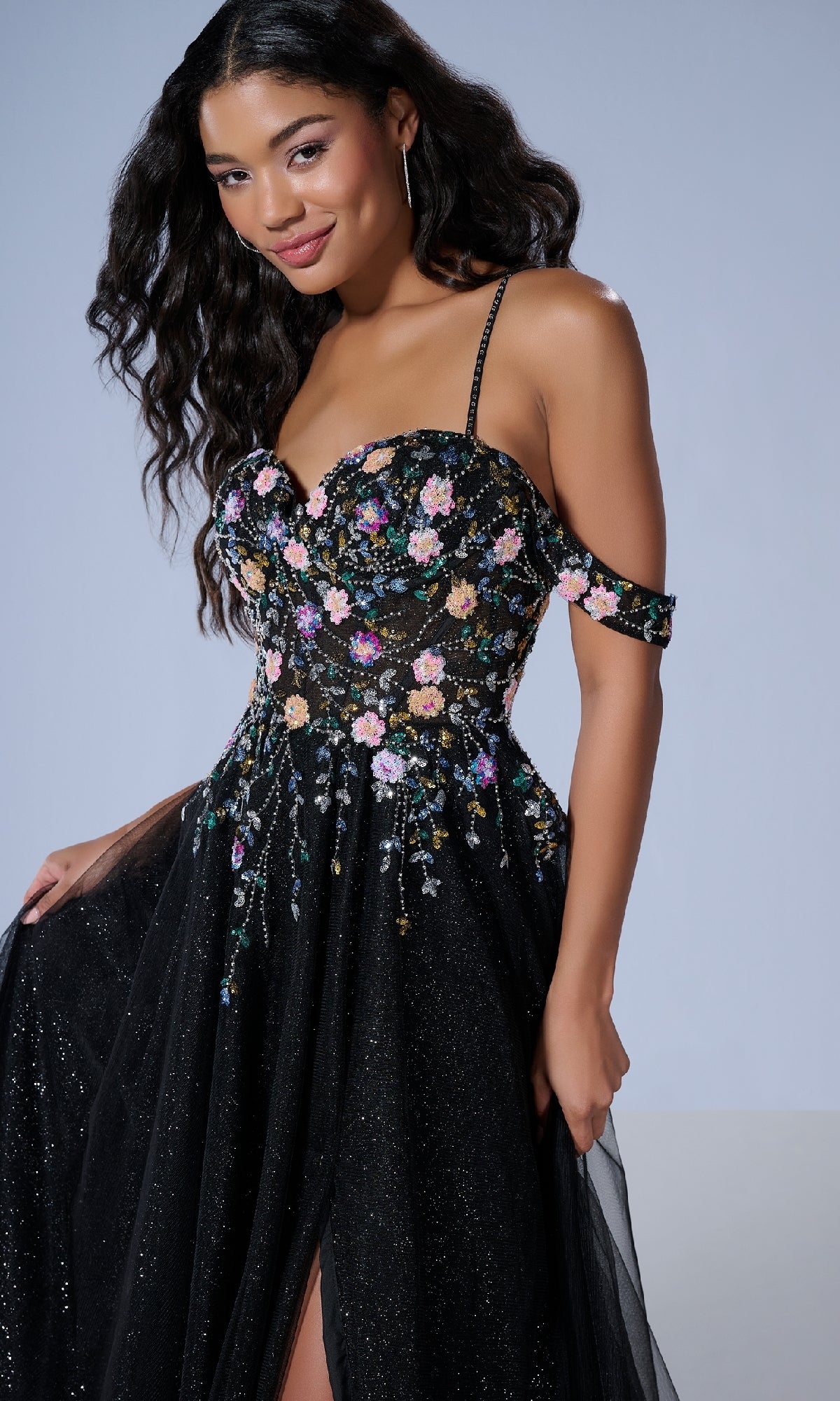 A woman stands against a plain background wearing the Tiffany 16251 long prom dress, a black off-shoulder A-line gown with colorful floral embroidery, thin straps, and delicate floral beaded appliqué.
